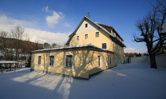 A yellow building stands in a snowy landscape. The sky is clear and blue, and the shadows of the trees are visible on the snow.