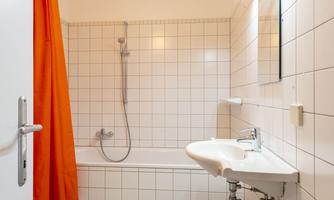 A modern bathroom with white tiles and a sink. The shower is integrated into the bathtub and there is a bright mirror.