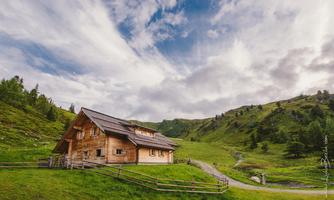An idyllic wooden house surrounded by green meadows and mountains. The sky is partly cloudy and the landscape appears peaceful.