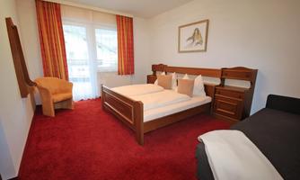 A cozy bedroom with a double bed and a single bed. The room is bright and friendly with red curtains and a chair in the corner.