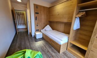 A simple room with a single bed and wooden furniture. In the background, a wardrobe and a green seat cushion can be seen.