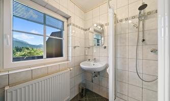 A modern bathroom with a shower, a sink, and a window that offers a view of the landscape. The walls are light tiled and the space feels inviting and clean.