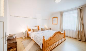 A bright bedroom with a large double bed and two pillows. Large windows with curtains let in plenty of light and create an inviting atmosphere.
