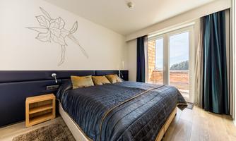 A modern bedroom with a large bed and a flower illustration on the wall. Large windows provide plenty of daylight and a view outside.