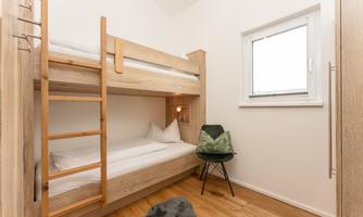 A simple bedroom with an unfolded wooden bunk bed. A cozy chair and a window provide daylight.