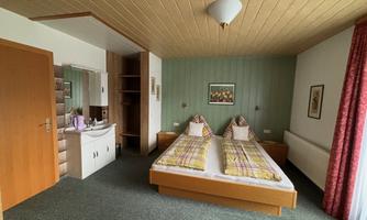A simple bedroom with two beds and a wooden wardrobe. A picture hangs on the wall, and the room has carpet flooring and windows with curtains.