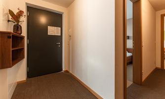 A bright hallway with a dark front door. A shelf hangs on the wall, and there is a connecting door to another room.