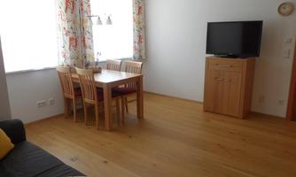 A bright living room with a wooden table and chairs. In the background, there is a television and large windows with floral curtains.
