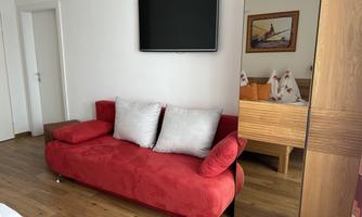 A modern living room with a red sofa and a flat-screen television on the wall. In the background, a wooden cabinet and a bed are visible.