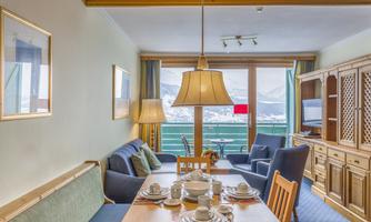 A cozy living room with a dining table, chairs, and a cabinet. Large windows offer a view of the mountains and the balcony.