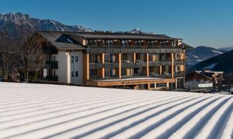 A modern building sits on a snow-covered field. In the background, mountains are visible under a clear blue sky.