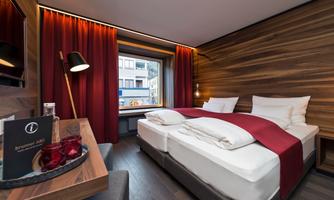 A modern hotel room with a cozy double bed and warm wood paneling. The large windows let in plenty of light, and the room is stylishly decorated with red curtains.