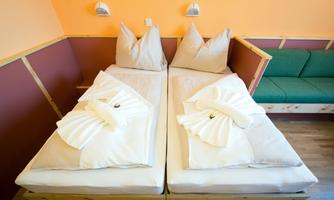 A cozy room with two single beds and fresh towels. The walls are painted in warm colors, and a comfortable sofa is visible.