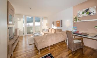 A modern living room with a cozy sofa and a television. Large windows offer a view of the surrounding mountains and plenty of daylight.