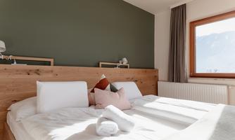 A cozy bedroom with a wooden headboard and colorful pillows. The window offers a view of nature and allows for plenty of daylight.