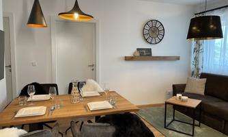 A modern dining room with a wooden table and stylish chairs. A clock hangs on the wall, and there is cozy lighting above the table.