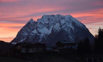 A majestic mountain with a snow-capped peak is illuminated by a colorful sunset. In the foreground, some houses and trees can be seen.