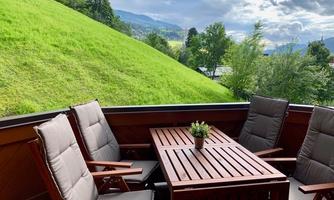 A cozy balcony with a wooden table and comfortable chairs. In the background, a green meadow and trees can be seen.