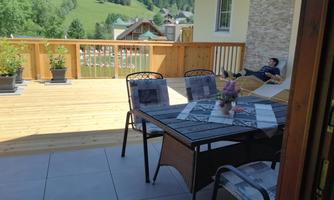 A cozy balcony with a rattan table and chairs. In the background, there are green hills and a relaxed person visible.