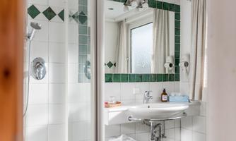 A modern bathroom with white tiles and green accents. 
It features a shower, a sink, and large windows that provide plenty of light.