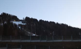 A snow-covered slope with trees and a clear blue sky in the background. In the foreground, a wall is visible.