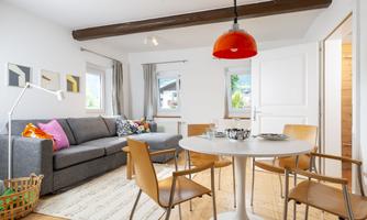 A modern living room with a gray sofa and a round dining table. The windows let in a lot of light and the walls are painted light.