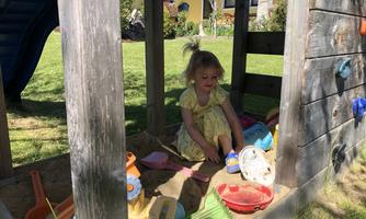 A little girl is playing in a wooden playhouse. Colorful sand shovels and toys are scattered around her.