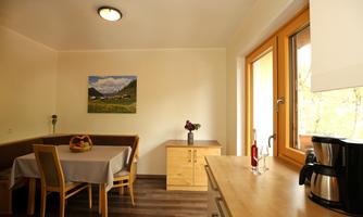 A bright and cozy kitchen with a dining table and chairs. A picture of a landscape hangs on the wall, and there are large windows that let in daylight.