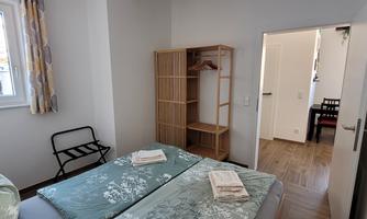A simple bedroom with two beds, a shelf, and a chair. The room is bright and friendly, with a window that lets in natural light.