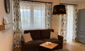 A cozy living room with a brown sofa and a small table. Large windows let in plenty of daylight and the curtains have a floral design.