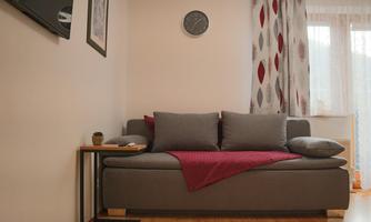 A cozy living room with a gray sofa and red cushions. Next to the sofa is a small table and a clock hangs on the wall.