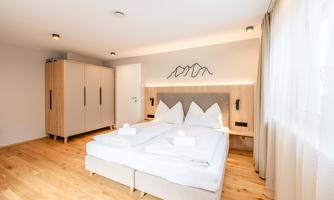 A modern bedroom with a double bed and wooden flooring. The walls are light and there is a large wardrobe as well as windows with curtains.