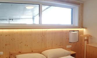 A cozy bedroom with wooden walls and a large window. The bed is equipped with beige pillows and blankets.