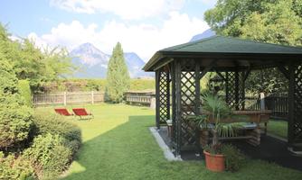 A beautiful garden with a pavilion, two loungers, and green plants. In the background, mountains and a blue sky can be seen.