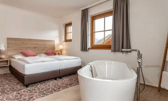 A modern bedroom with a large bed and a freestanding bathtub. Large windows let in plenty of daylight and offer a view outside.