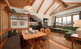 A cozy living space with wooden furniture and large windows that offer a view of the mountains. The room is bright and inviting, featuring a dining table and a sofa area.