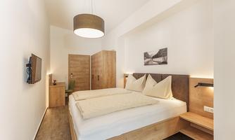 A cozy bedroom with a double bed and modern wooden furniture. The walls are light, and there is a large lampshade as well as a television.
