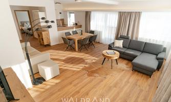 A modern living room with a gray sofa and a round table. The room is bright and cheerful with large windows and wooden flooring.