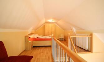 A cozy attic room with a bed and a red seating area. Warm light and wooden flooring create an inviting atmosphere.
