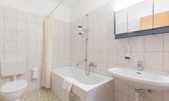 A bright bathroom with a bathtub, shower, and sink. The room is fitted with white tiles and a large mirror.