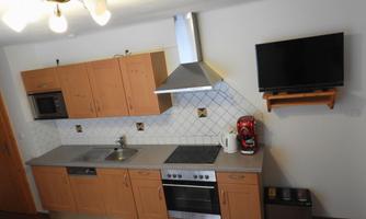 A modern kitchen with wooden cabinets, a stove, and a range hood. A television is mounted on the wall, and there is a sink as well as a coffee machine.