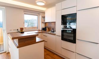 A modern kitchen with white cabinets and wooden details. There are large windows that let in plenty of light.