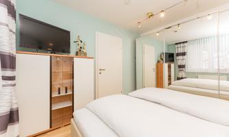 A modernly furnished bedroom with a double bed, a television on the wall, and a wardrobe. The walls are painted in a soft green tone, and large windows let in plenty of daylight.