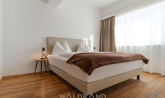 A modern bedroom with a cozy bed and a large window front. The wooden floors and the bright design create an inviting atmosphere.
