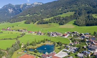 A picturesque landscape with a small pond and green meadows. In the background, mountains and a charming village can be seen.