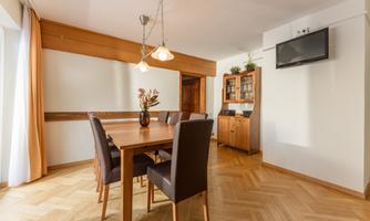 A bright dining area with a wooden table and elegant chairs. There is a television and a showcase on the wall.