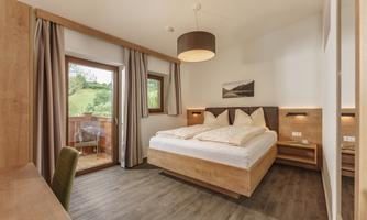 A modern bedroom with a large bed and wooden decor. There are windows overlooking greenery and a desk.