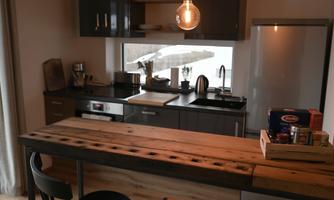A modern kitchen with a wooden countertop and black bar stools. The room feels bright and inviting, with large windows and minimally furnished cabinets.