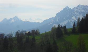 A picturesque landscape with green meadows and snow-capped mountains in the background. A small farmhouse is situated on the slope, surrounded by trees.
