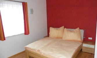 A simple bedroom with a double bed and bright pillows. The walls are painted in warm tones and a window provides natural light.